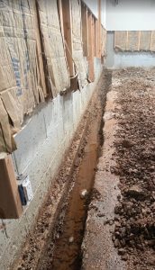 Basement showing trench for French drain installation, with exposed concrete walls and damp soil, indicating moisture issues and need for waterproofing solutions.