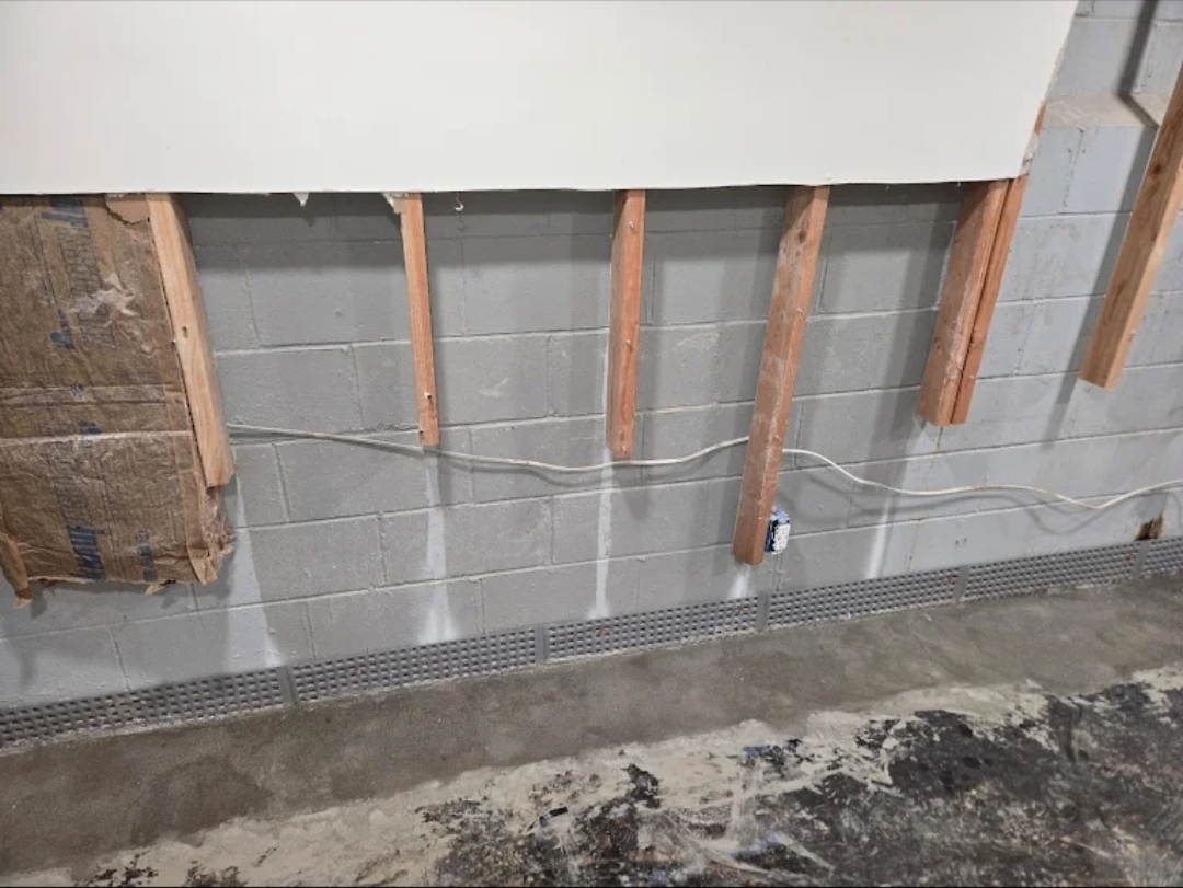 Interior basement drainage system installed along foundation wall with visible moisture marks and framing exposed